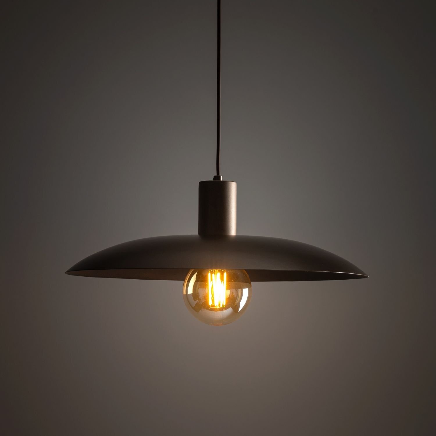 Na obrazie widoczna jest brązowa lampa wisząca z metalowym kloszem i żarówką emitującą ciepłe światło. Lampa zawieszona jest na czarnym przewodzie, a tło jest ciemne.