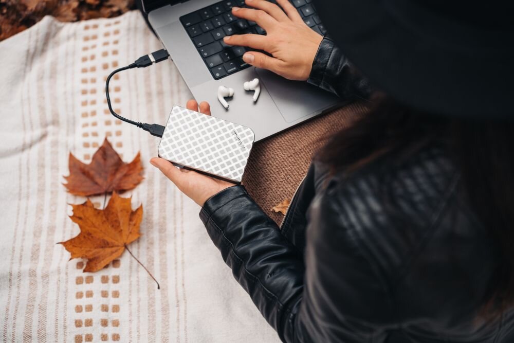 Dysk VERBATIM Store n Go 1TB HDD Srebrny Osoba w czarnej kurtce i kapeluszu trzyma w dłoni srebrny dysk z geometrycznym wzorem podłączony kablem do laptopa stojącego na kocu, obok leżą słuchawki bezprzewodowe i liście klonu, opis