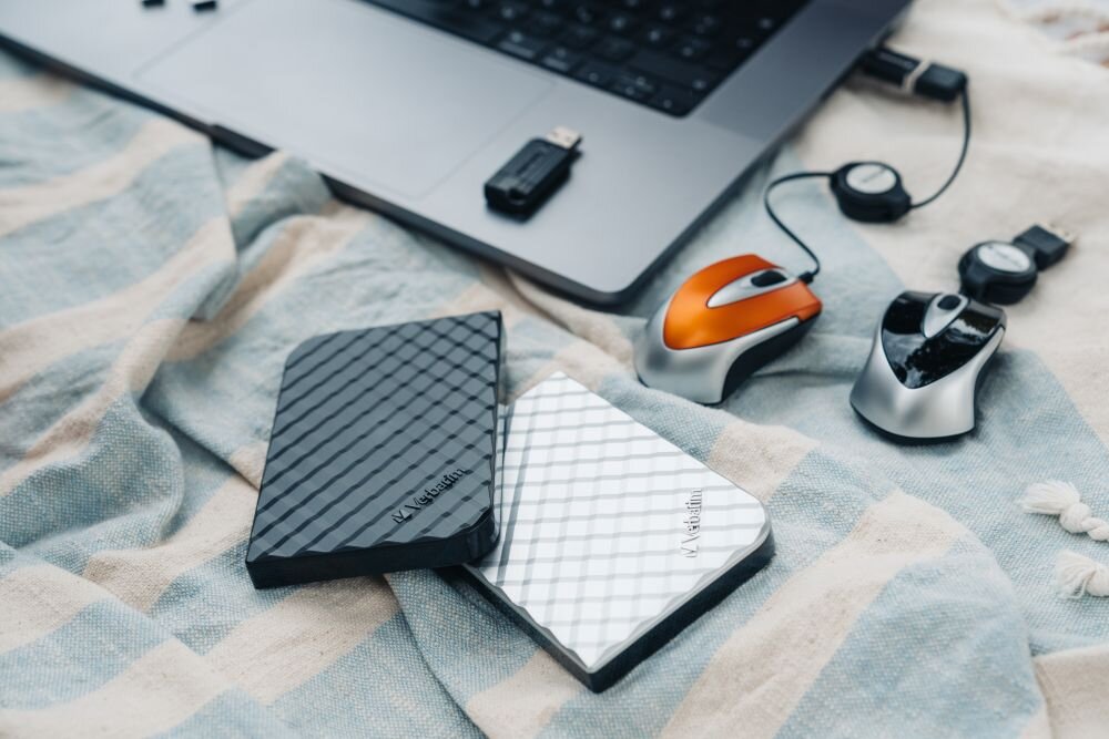 Dysk VERBATIM Store n Go 1TB HDD Srebrny Dwa dyski przenośne w kolorze czarnym i srebrnym leżące na materiale obok laptopa, dwóch przewodowych myszek i pendrive'a