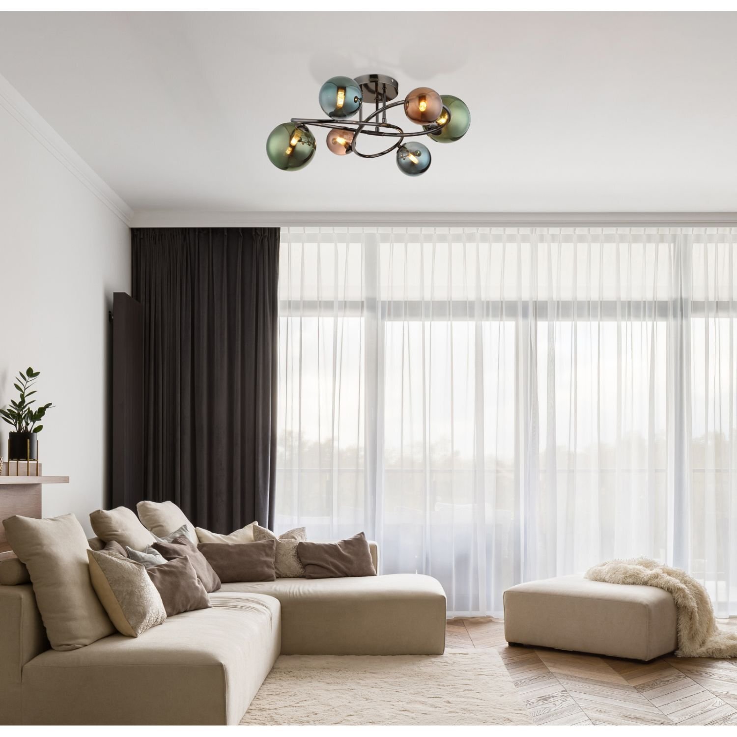 W salonie znajduje się nowoczesna lampa sufitowa z kolorowymi kloszami, zamontowana na suficie. W pomieszczeniu jest jasna sofa z poduszkami, a przez duże okna wpada naturalne światło.