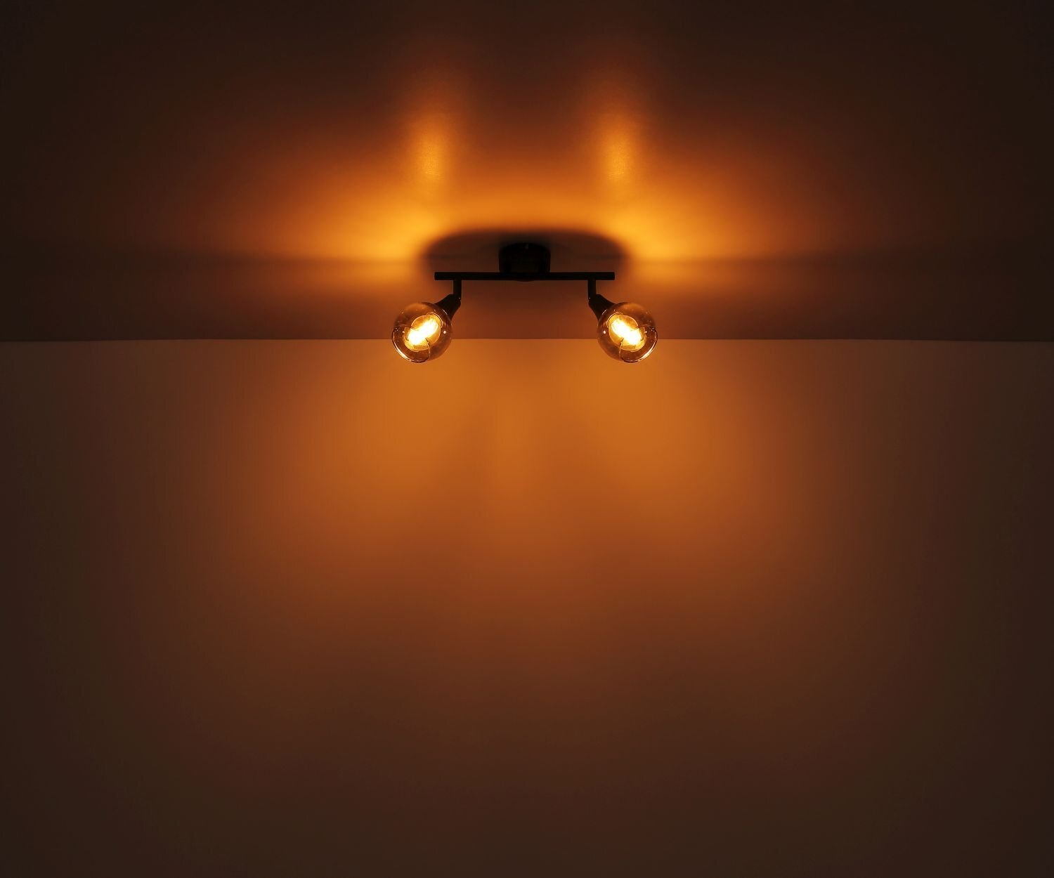 Na suficie zamontowana jest lampa sufitowa z dwoma reflektorami, emitująca ciepłe, żółte światło. Światło tworzy na suficie i ścianie delikatne, rozproszone refleksy.