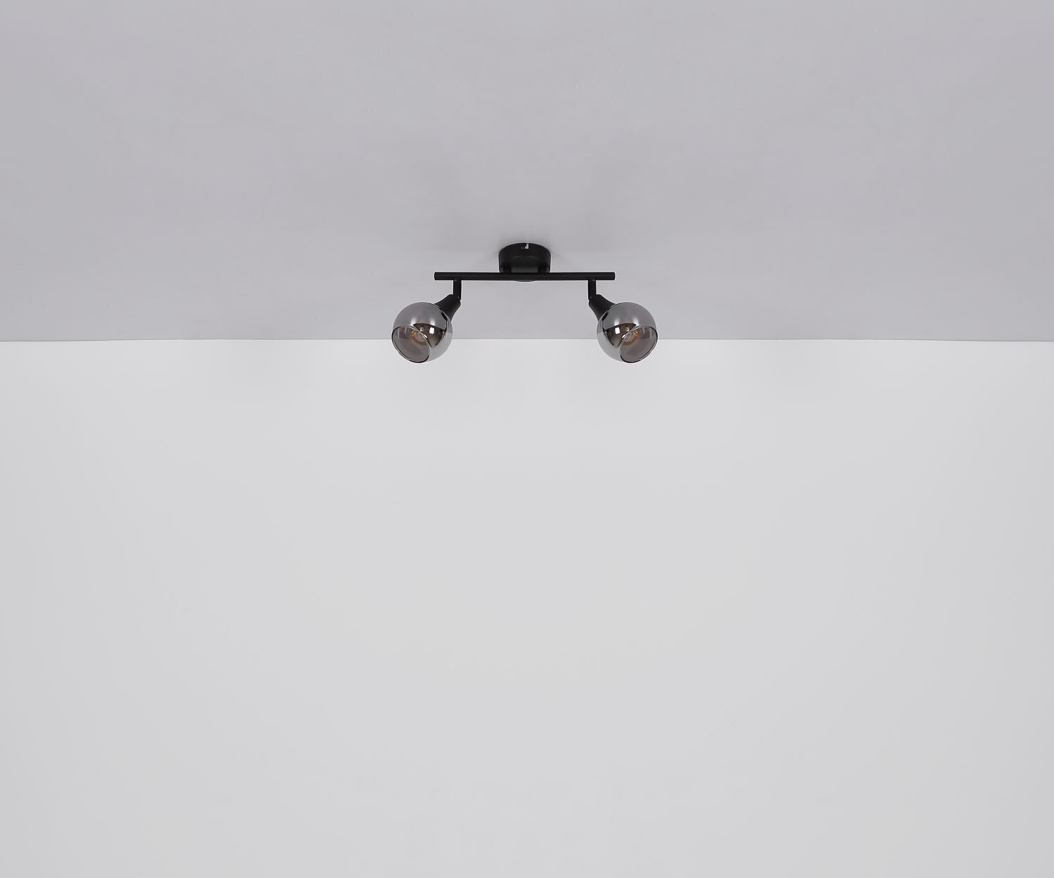 Na obrazie widoczna jest czarna lampa sufitowa z dwoma reflektorami zamontowana na białym suficie. Reflektory mają kulisty kształt i są skierowane w dół.