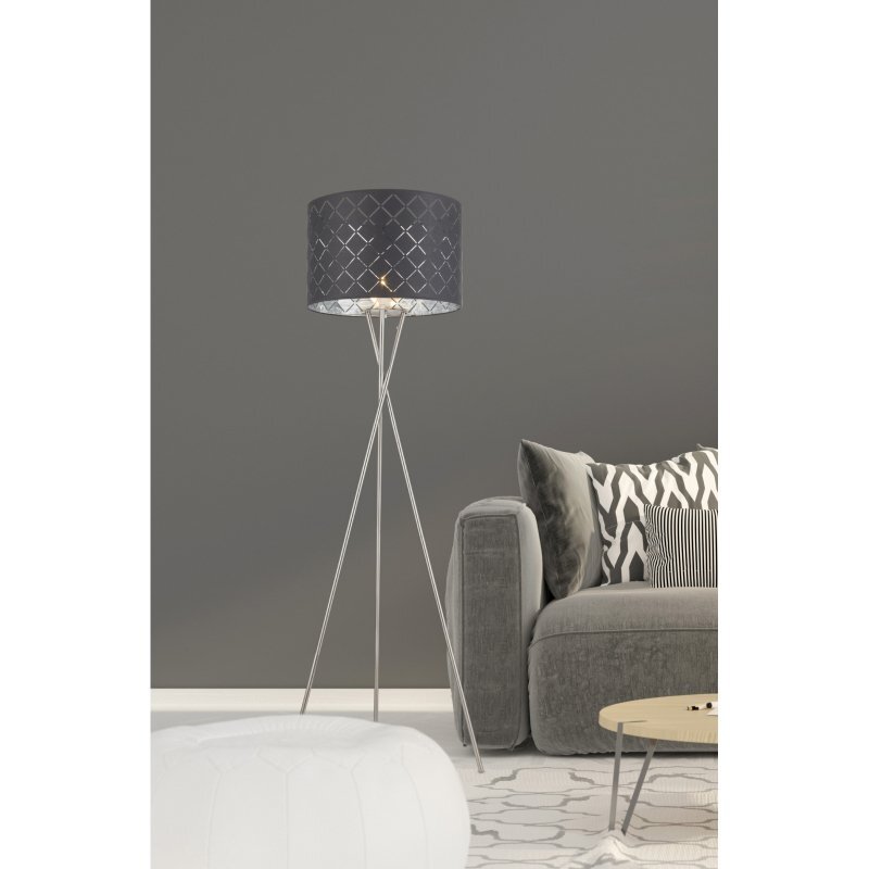 Na obrazie znajduje się lampa podłogowa z szarym tekstylnym abażurem na trójnogu, ustawiona obok szarej kanapy z poduszkami w czarno-białe wzory. W tle widoczna jest szara ściana, a na podłodze znajduje się jasny dywan z geometrycznym wzorem.