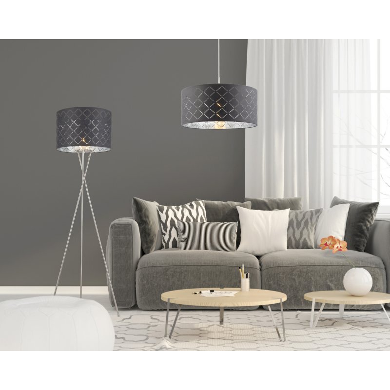 W salonie znajduje się szara sofa z różnorodnymi poduszkami, obok której stoi lampa podłogowa z szarym abażurem. Na stolikach kawowych leżą drobne przedmioty, a w tle widać zasłony i okno.