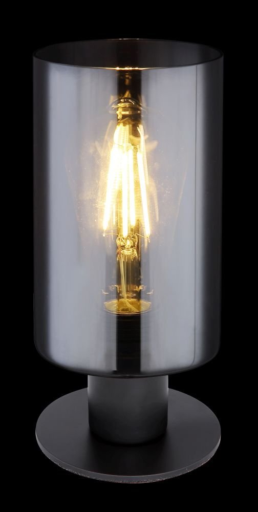 Czarna matowa lampa stołowa z przydymionym szklanym kloszem, przez który widać żarówkę emitującą ciepłe światło. Lampa stoi na okrągłej podstawie.