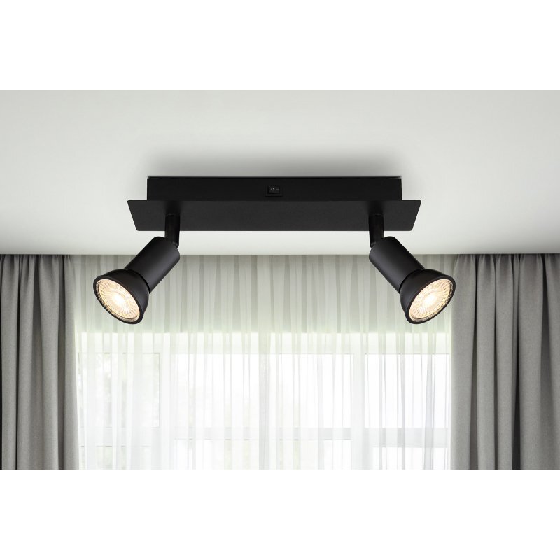 Czarna matowa lampa sufitowa z dwoma reflektorami skierowanymi w dół, zamontowana na prostokątnej podstawie. W tle widoczne są zasłony i okno z białą firanką.