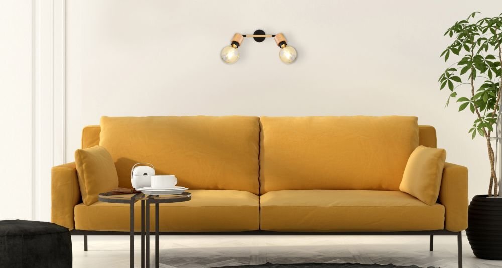Na ścianie nad musztardową sofą zamontowana jest lampa ścienna z dwoma żarówkami, wykonana z czarnego matowego metalu i drewna. Obok sofy stoi stolik z filiżanką i talerzykiem, a w rogu znajduje się roślina w doniczce.