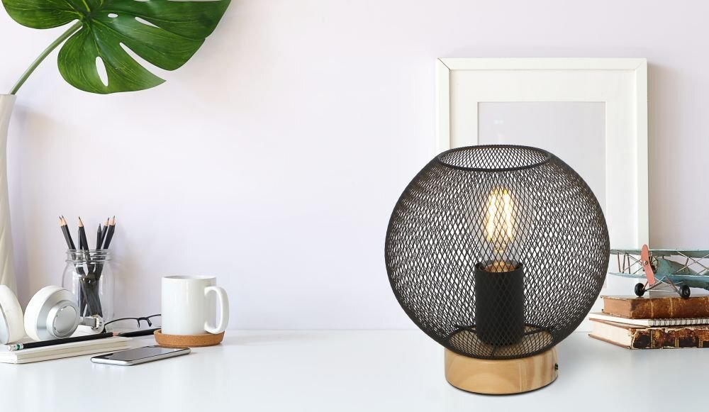 Na biurku stoi lampa stołowa z czarnym, ażurowym kloszem i drewnianą podstawą. Obok lampy znajdują się książki, model samolotu, kubek, słuchawki i roślina w doniczce.