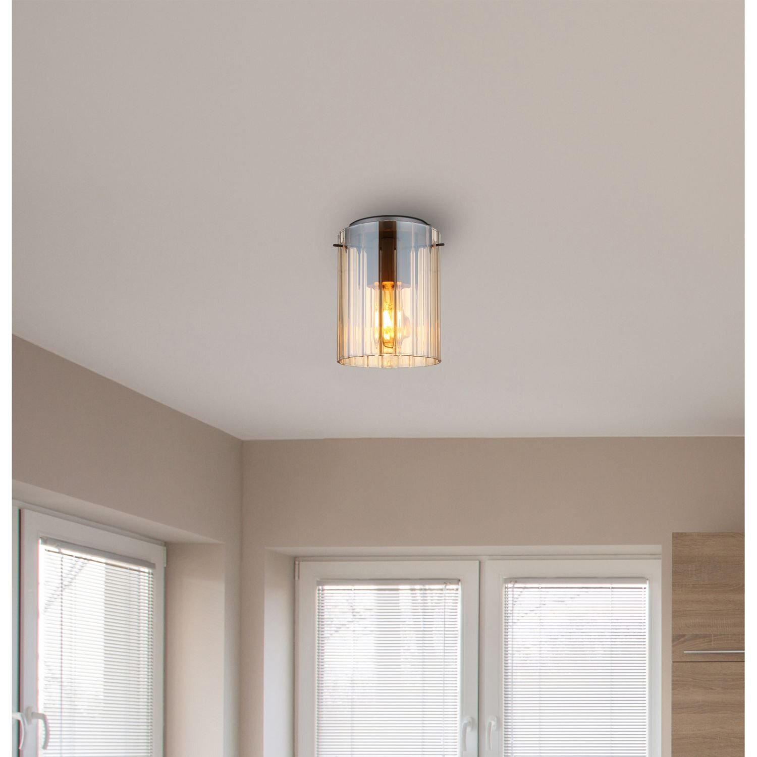 Na suficie zamontowana jest lampa z ryflowanym kloszem, przez który widać żarówkę. W tle widoczne są okna z żaluzjami i fragment ściany.