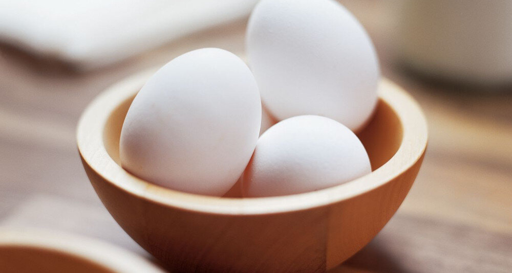 Trzy białe jajka w drewnianej misce na rozmytym tle.