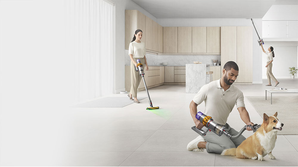 Na obrazie widać trzy osoby używające odkurzacza bezprzewodowego w nowoczesnym wnętrzu kuchennym. Jedna osoba siedzi na podłodze obok psa, trzymając odkurzacz.