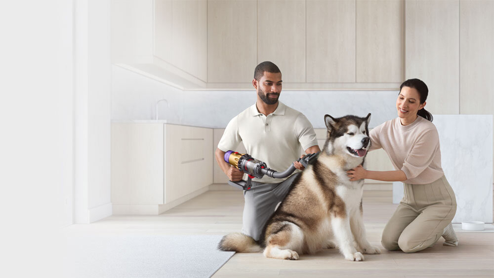 Dwie osoby znajdują się w jasnym pomieszczeniu, jedna z nich trzyma odkurzacz, a obok nich siedzi duży pies. Odkurzacz ma nowoczesny design i jest używany do pielęgnacji sierści psa.