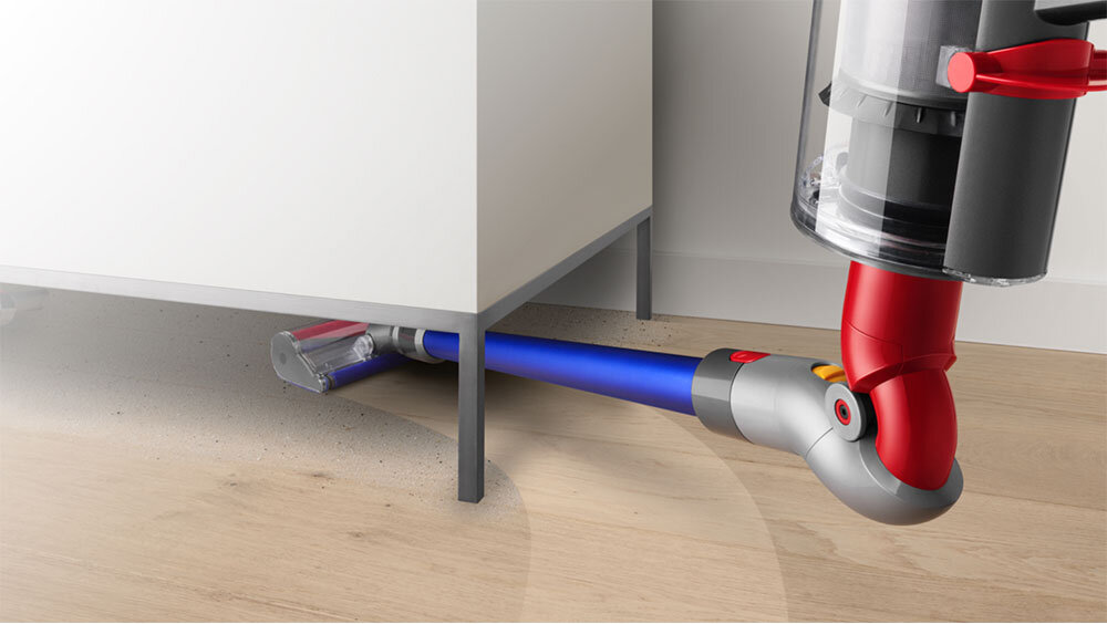 Odkurzacz z niebieską rurą i przezroczystym pojemnikiem na kurz odkurza podłogę pod białą szafką. Na podłodze widoczne są drobinki kurzu.