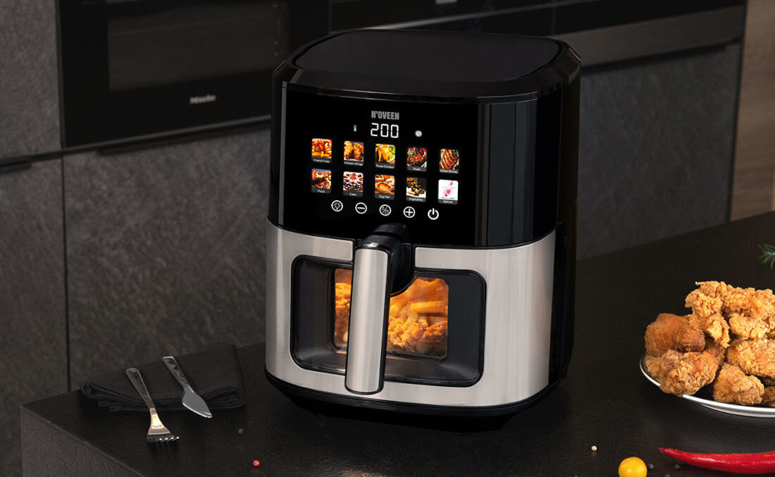 Na blacie kuchennym stoi frytkownica beztłuszczowa z wyświetlaczem pokazującym temperaturę 200 stopni oraz różne ikony programów. Obok frytkownicy znajduje się talerz z kawałkami smażonego kurczaka, a na blacie leżą sztućce.