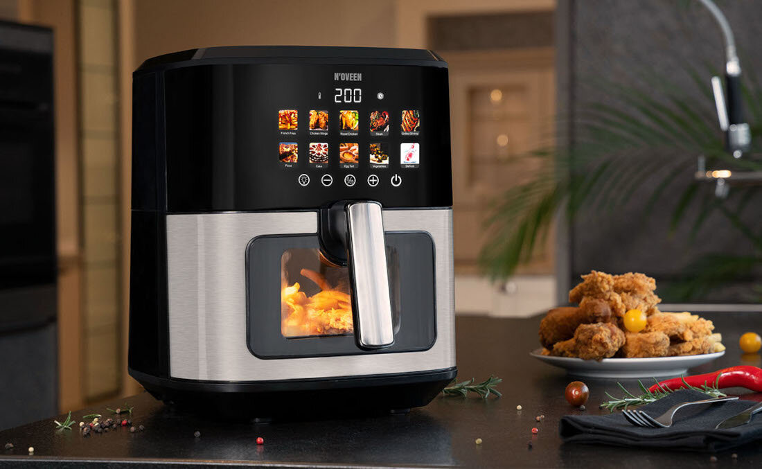 Na blacie kuchennym stoi frytkownica beztłuszczowa z wyświetlaczem pokazującym temperaturę 200 stopni oraz różne ikony programów. Obok znajduje się talerz z kawałkami smażonego kurczaka i pomidorkami koktajlowymi.