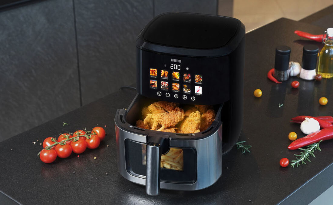 Na blacie kuchennym stoi frytkownica beztłuszczowa z wyświetlaczem pokazującym temperaturę 200 stopni i różne ikony programów. Wokół urządzenia znajdują się pomidory, papryczki i przyprawy.