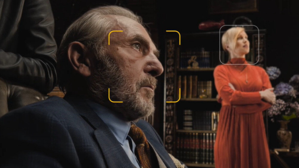 Na obrazie widać starszego mężczyznę w garniturze oraz kobietę w czerwonej sukni stojącą na tle półki z książkami. Na twarzach obu osób znajdują się żółte i białe ramki, sugerujące użycie funkcji rozpoznawania twarzy.
