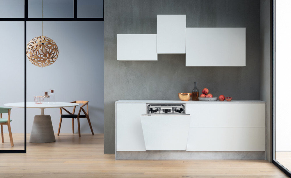 Nowoczesna kuchnia z otwartą zmywarką w zabudowie, obok której znajdują się białe szafki i blat z owocami. W tle widoczny jest stół z krzesłami oraz dekoracyjna lampa sufitowa.