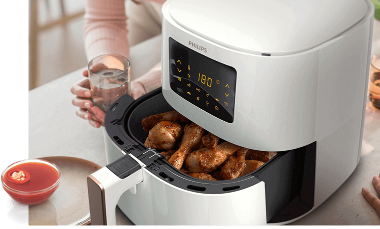 Biała frytkownica beztłuszczowa z wyświetlaczem pokazującym temperaturę 180 stopni i ikonami dotykowymi. W środku znajdują się upieczone kawałki kurczaka, a obok stoi szklanka z napojem.