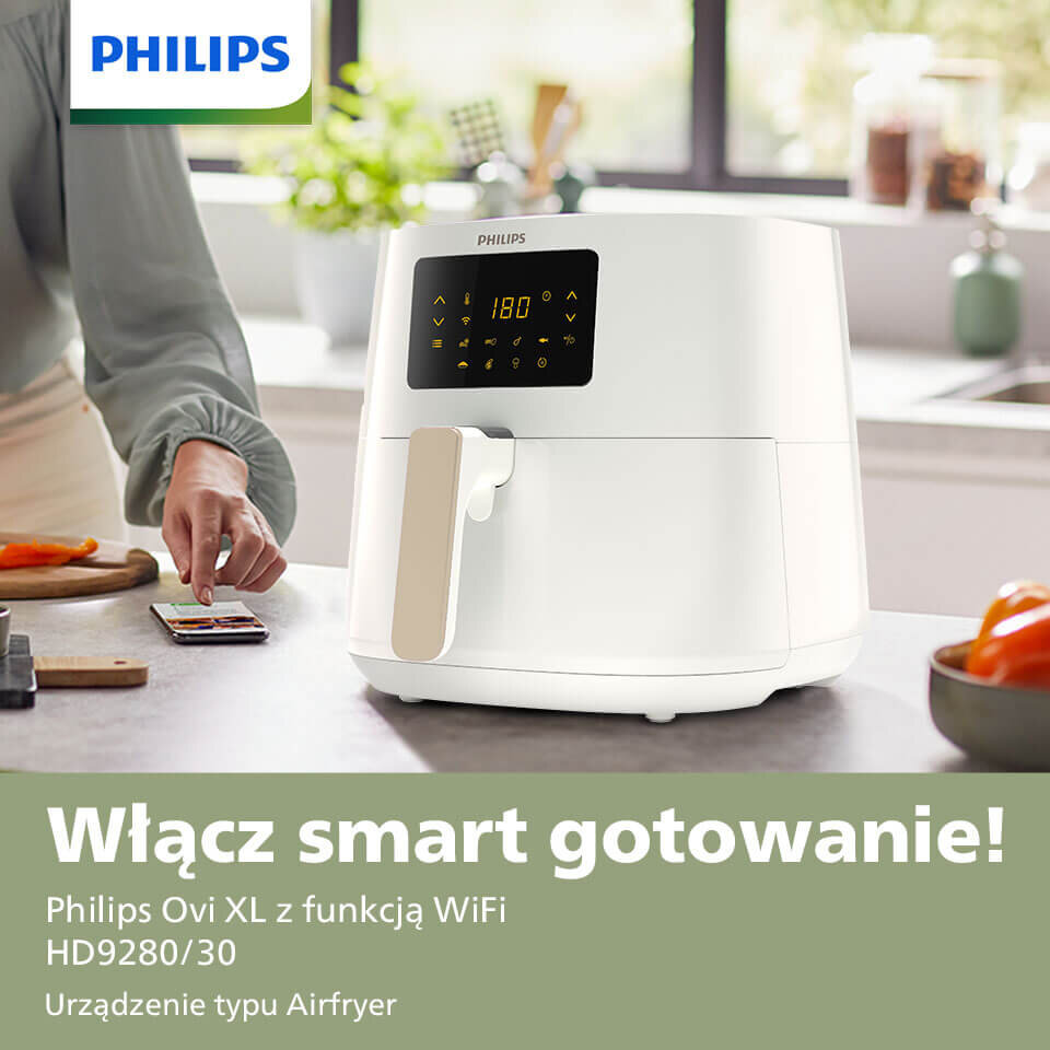 Na blacie kuchennym stoi biała frytkownica beztłuszczowa z wyświetlaczem pokazującym 180 stopni. Obok osoba obsługuje smartfon, a w tle widoczne są kuchenne akcesoria i warzywa.