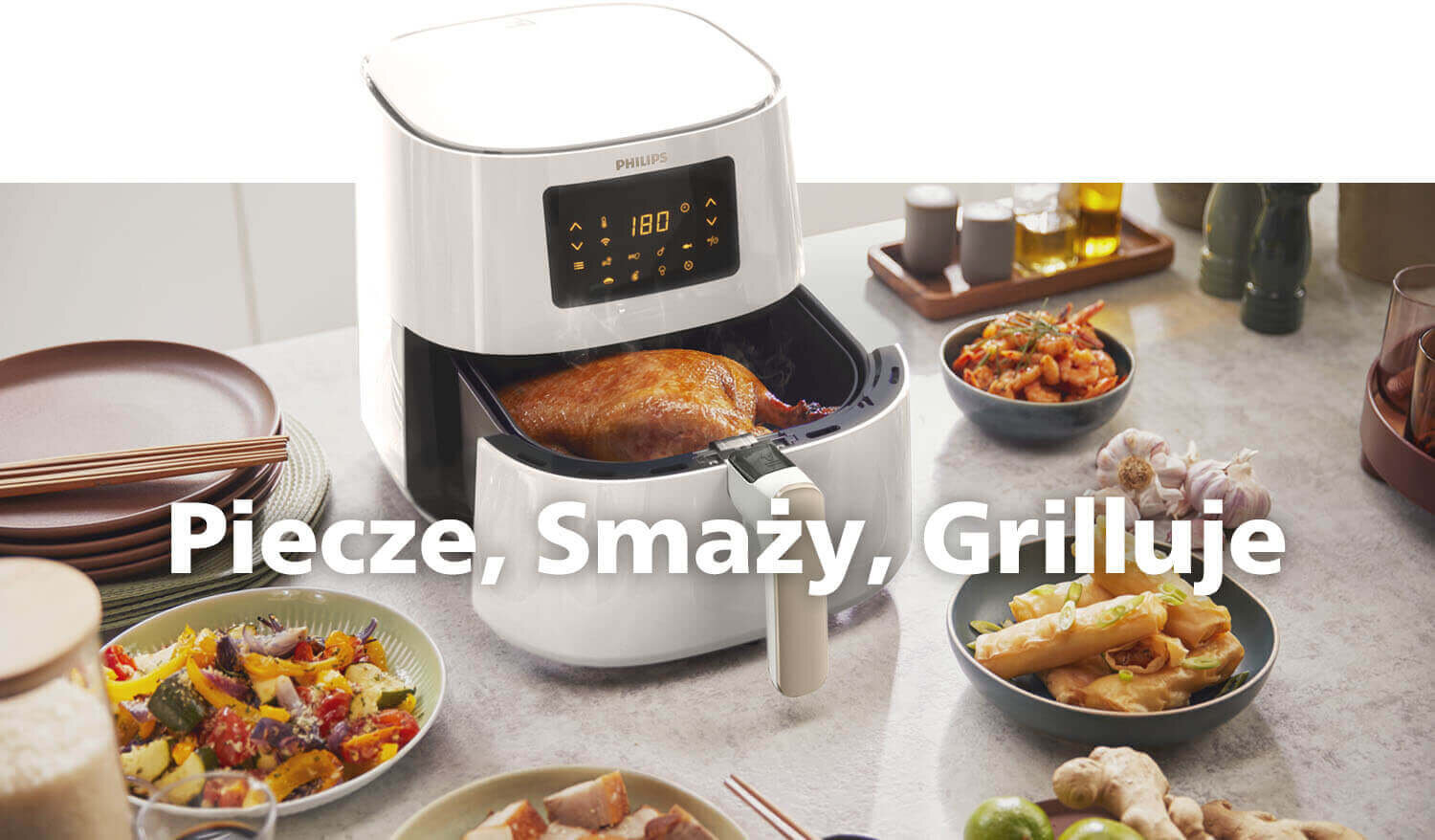 Na blacie kuchennym stoi biała frytkownica beztłuszczowa z wyświetlaczem pokazującym 180°C, w której znajduje się pieczony kurczak. Wokół urządzenia znajdują się różne potrawy i przyprawy, a na obrazie widnieje tekst: \'Piecz, Smaż, Grilluj\'.