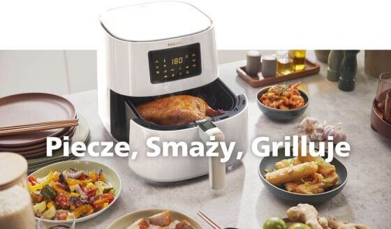 Na blacie kuchennym stoi biała frytkownica beztłuszczowa z wyświetlaczem pokazującym 180 stopni, w której znajduje się pieczony kurczak. Wokół frytkownicy znajdują się różne potrawy i przyprawy, a na obrazie widnieje tekst: \'Pieczę, Smaży, Grilluje\'.