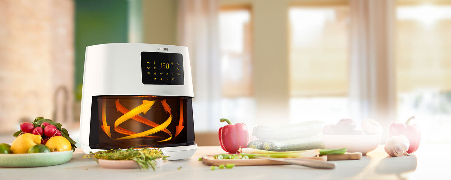 Na blacie kuchennym stoi biała frytkownica beztłuszczowa z wyświetlaczem pokazującym temperaturę 180°C. Obok znajdują się różne warzywa, w tym papryka i cytryna.
