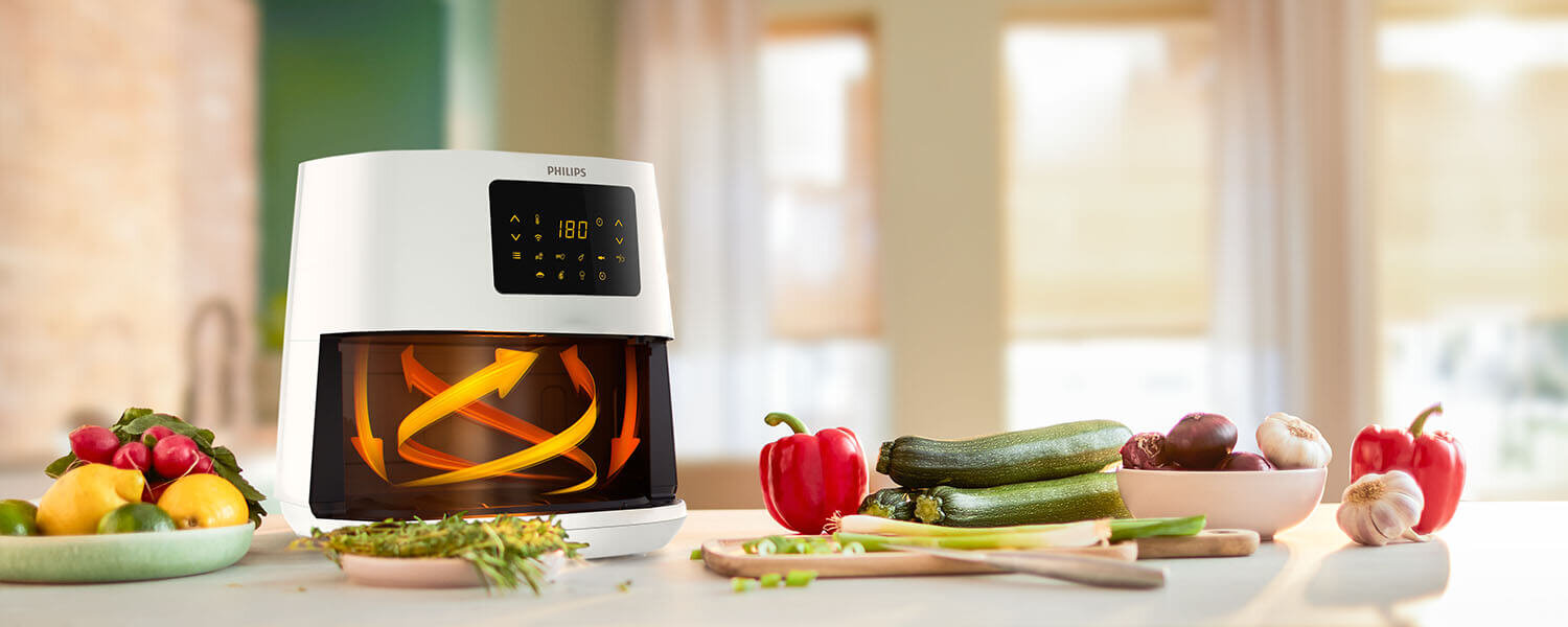 Na blacie kuchennym stoi biała frytkownica beztłuszczowa z wyświetlaczem pokazującym temperaturę 180°C. Obok znajdują się różne warzywa, takie jak papryka, cukinia i cebula.