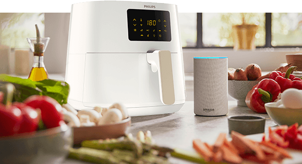 Na blacie kuchennym stoi biała frytkownica beztłuszczowa z wyświetlaczem pokazującym temperaturę 180°C. Obok niej znajduje się głośnik z napisem \'amazon\' oraz różne warzywa, takie jak papryki i pieczarki.