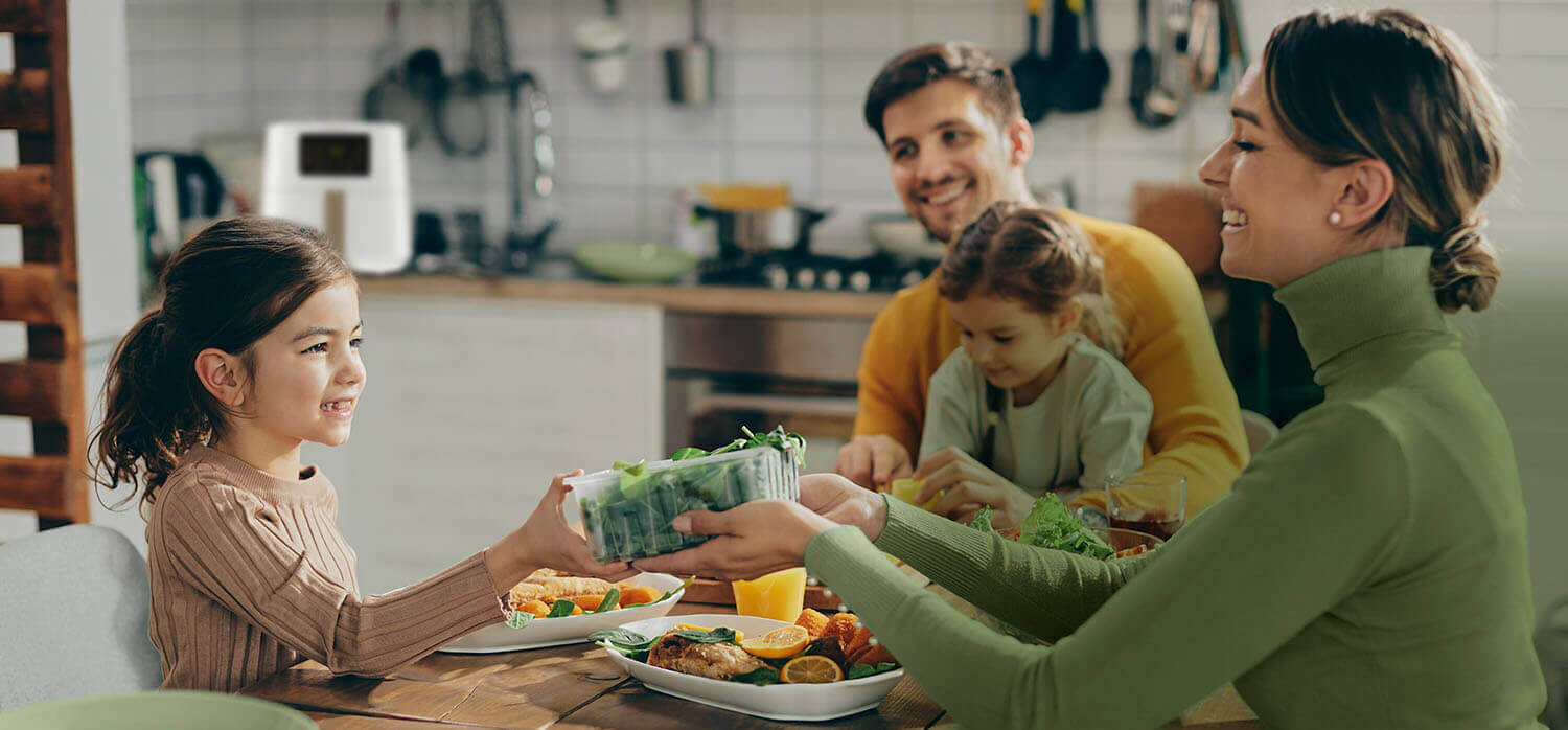 Rodzina siedzi przy stole w kuchni, na którym znajdują się różne potrawy. W tle widoczna jest frytkownica beztłuszczowa na blacie kuchennym.