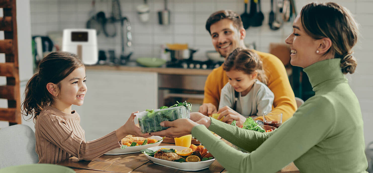 Rodzina siedzi przy stole w kuchni, na którym znajdują się talerze z jedzeniem. W tle widoczna jest frytkownica beztłuszczowa na blacie kuchennym.
