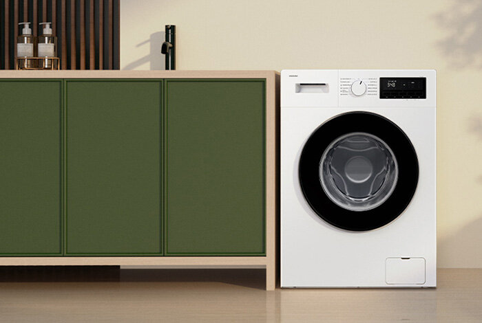 Biała pralka z czarnym panelem sterowania i okrągłym bębnem, ustawiona obok zielonej szafki. Na panelu widoczny jest napis \'SAMSUNG\'.