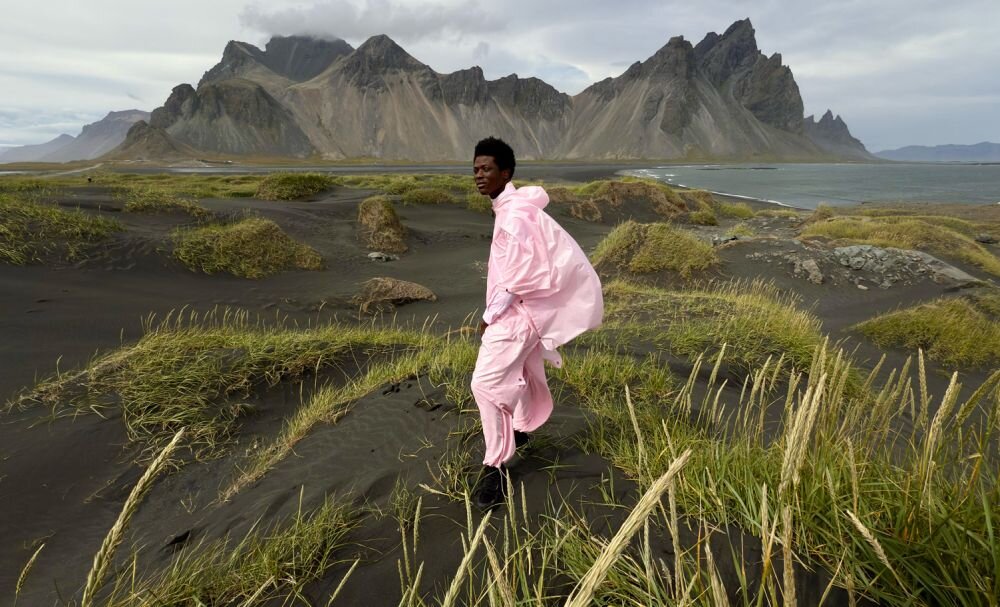 Osoba ubrana w różowy strój stoi na tle krajobrazu z czarnym piaskiem, trawą i górami w oddali. Niebo jest zachmurzone, a w tle widać wodę.