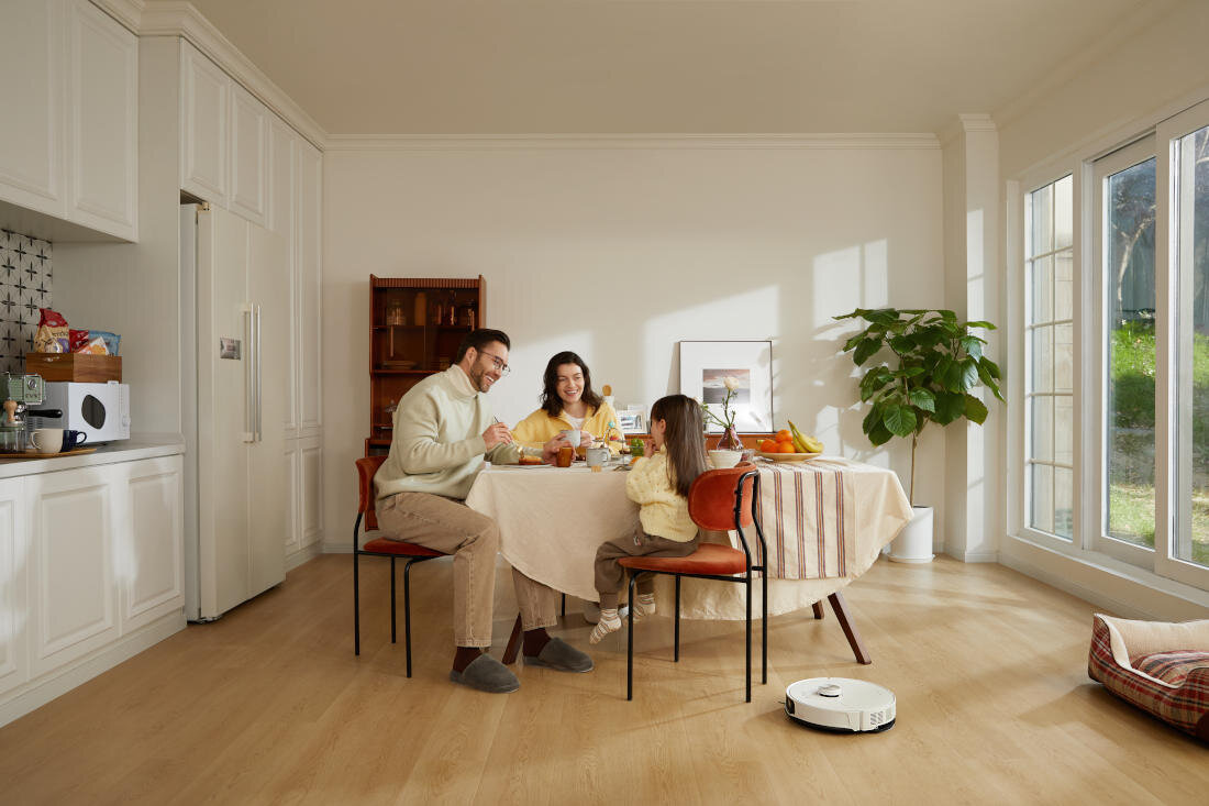 W jasnym pokoju z dużymi oknami, przy okrągłym stole siedzą trzy osoby, a na podłodze znajduje się biały robot sprzątający. W tle widać roślinę doniczkową i drewniany regał.