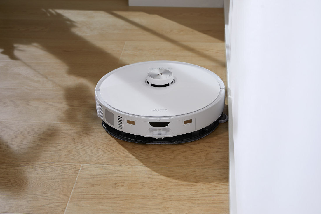 Biały robot sprzątający porusza się po drewnianej podłodze w pobliżu ściany. Na górze urządzenia widoczne jest logo producenta.