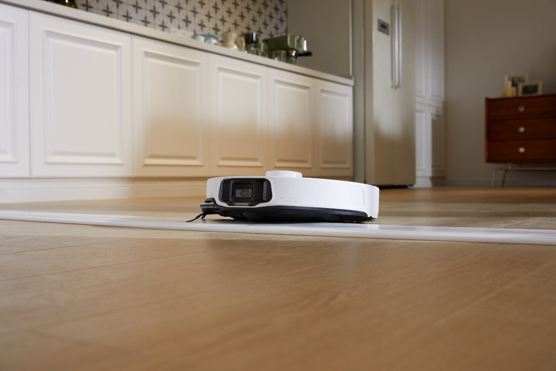 Na drewnianej podłodze w kuchni znajduje się biały robot sprzątający. W tle widoczne są białe szafki kuchenne i lodówka.
