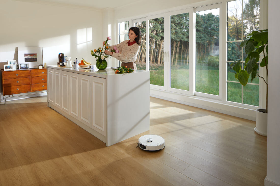 W jasnym pomieszczeniu z dużymi oknami znajduje się kobieta przy wyspie kuchennej, układająca kwiaty w wazonie. Na podłodze widoczny jest biały robot sprzątający.