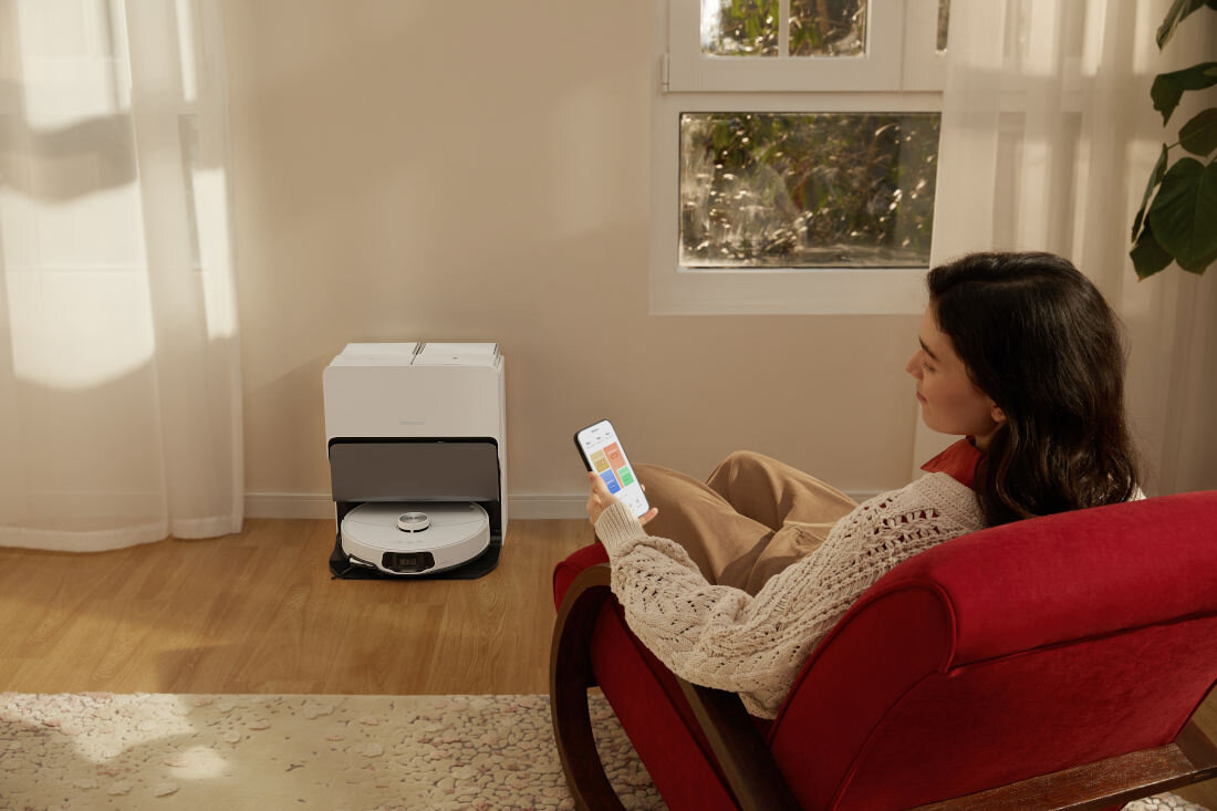 Osoba siedzi na czerwonym fotelu, trzymając w ręku smartfon z kolorowym ekranem. Obok znajduje się biały robot sprzątający w stacji dokującej na drewnianej podłodze.