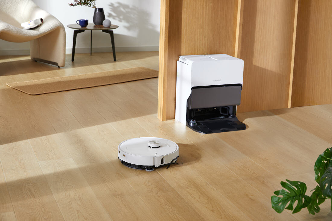 Na drewnianej podłodze znajduje się biały robot sprzątający oraz stacja dokująca. W tle widoczny jest stolik z wazonem i krzesło.