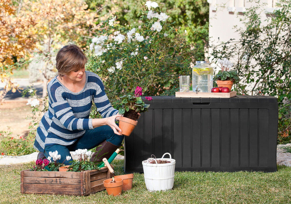 Osoba siedzi na trawie obok antracytowej skrzyni ogrodowej, trzymając doniczkę z rośliną. Na skrzyni stoją szklany dzbanek z napojem, szklanka oraz doniczka z rośliną.