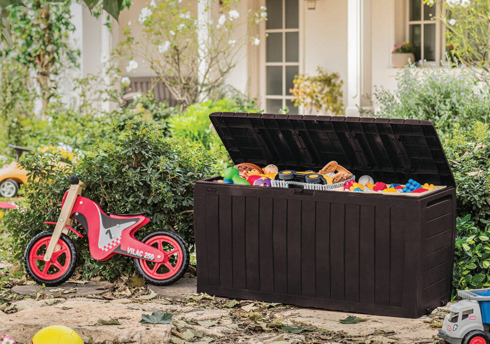 Na obrazie widoczna jest otwarta skrzynia ogrodowa wypełniona kolorowymi zabawkami, stojąca na tle zieleni. Obok skrzyni znajduje się czerwony rowerek z napisem \'VILAC 250\'.