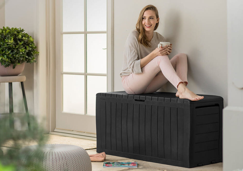 Osoba siedzi na antracytowej skrzyni ogrodowej, trzymając kubek. W tle widać jasną ścianę i okno.