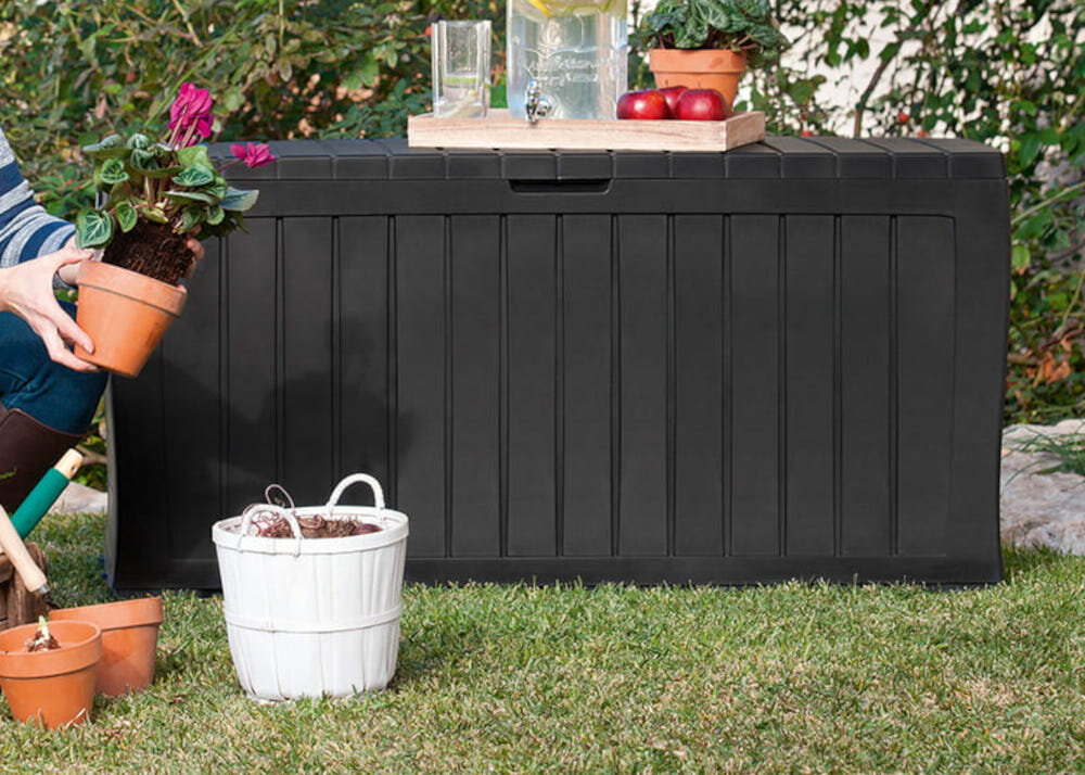 Na trawie stoi antracytowa skrzynia ogrodowa z pionowymi żłobieniami. Na skrzyni znajduje się taca z napojami i owocami, a obok niej widoczne są doniczki z roślinami.