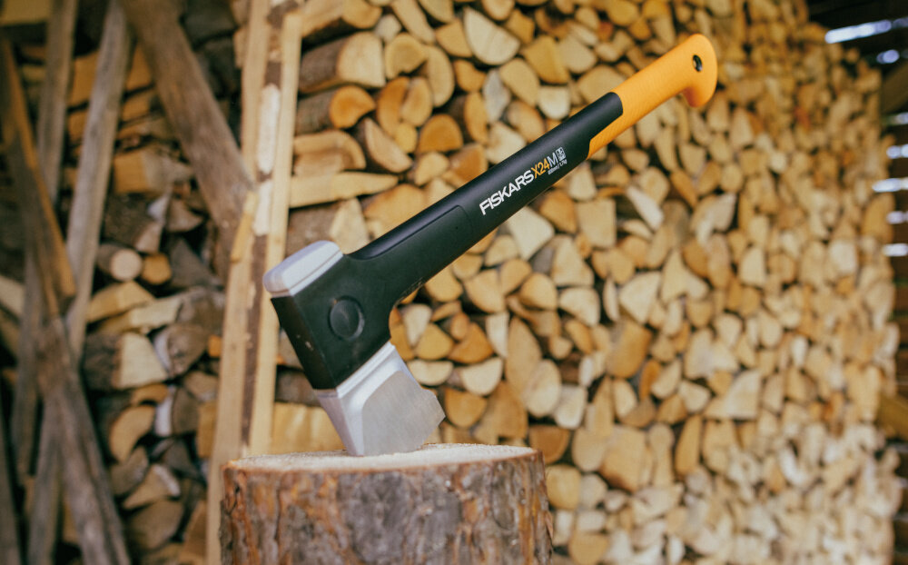 Siekiera z czarnym trzonkiem i pomarańczowym uchwytem wbita w pień, w tle stos drewna. Na trzonku widoczny napis: \'FISKARS X24\'.