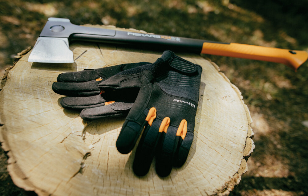 Na pniu drzewa leży siekiera z napisem \'FISKARS X24\' oraz para czarnych rękawic z pomarańczowymi akcentami i napisem \'FISKARS\'.