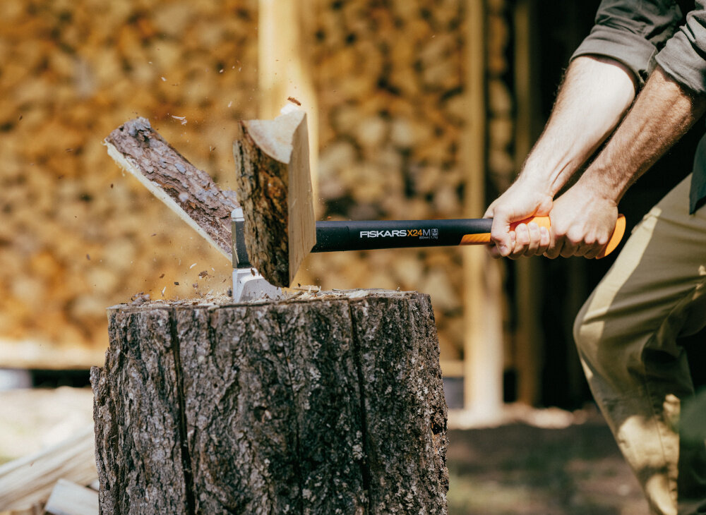 Osoba używa siekiery do rozłupywania drewna na pniu. Na trzonku siekiery widoczny jest napis \'FISKARS X24 M\'.