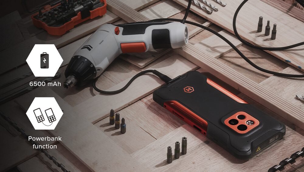 Na drewnianej powierzchni leży czarno-pomarańczowy smartfon z logo, obok którego znajduje się wkrętarka. Na obrazie widoczne są ikony z tekstem: \'6500 mAh\' oraz \'Powerbank function\'.