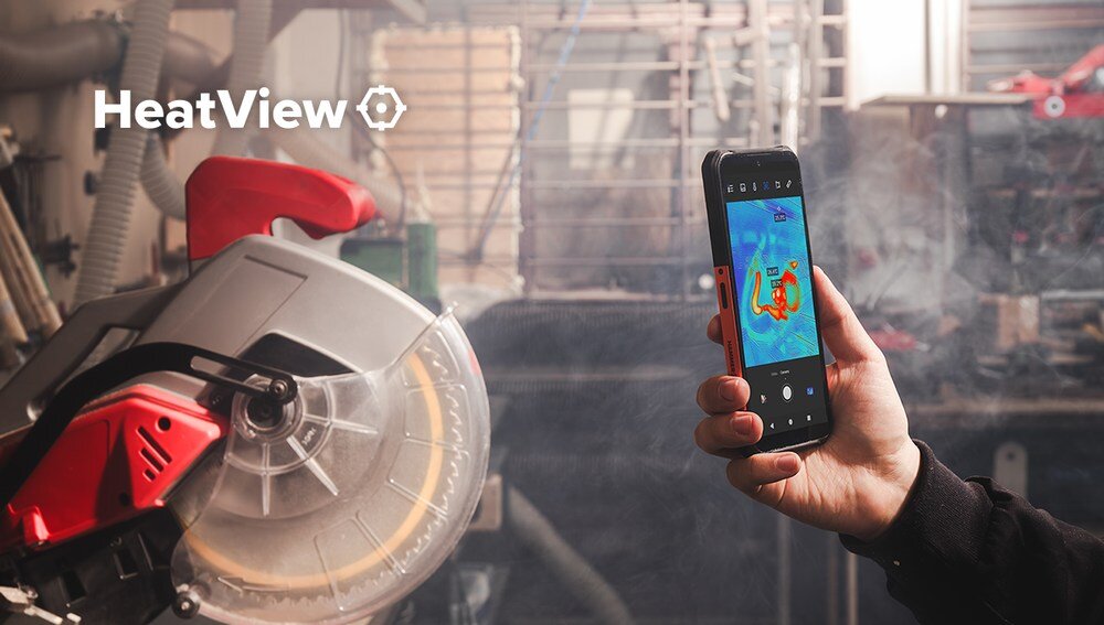 Osoba trzyma smartfon z włączoną aplikacją termowizyjną, na ekranie widoczny jest kolorowy obraz termiczny. W tle znajduje się piła tarczowa, a w lewym górnym rogu widnieje napis \'HeatView\'.