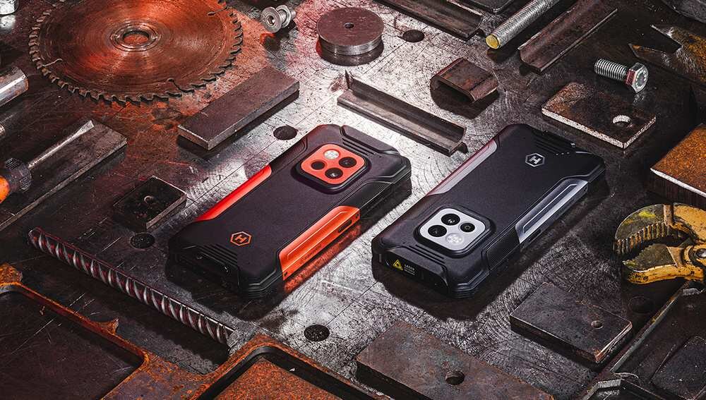 Dwa smartfony o solidnej konstrukcji leżą na metalowym stole w otoczeniu narzędzi i elementów mechanicznych. Obudowy telefonów są czarno-pomarańczowe i czarno-szare, z widocznym logo producenta.