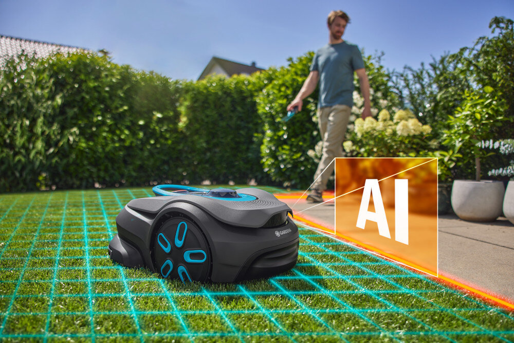 Na trawniku znajduje się robot koszący z widocznym logo \'GARDENA\', a obok niego widoczny jest napis \'AI\'. W tle widać osobę oraz żywopłot.